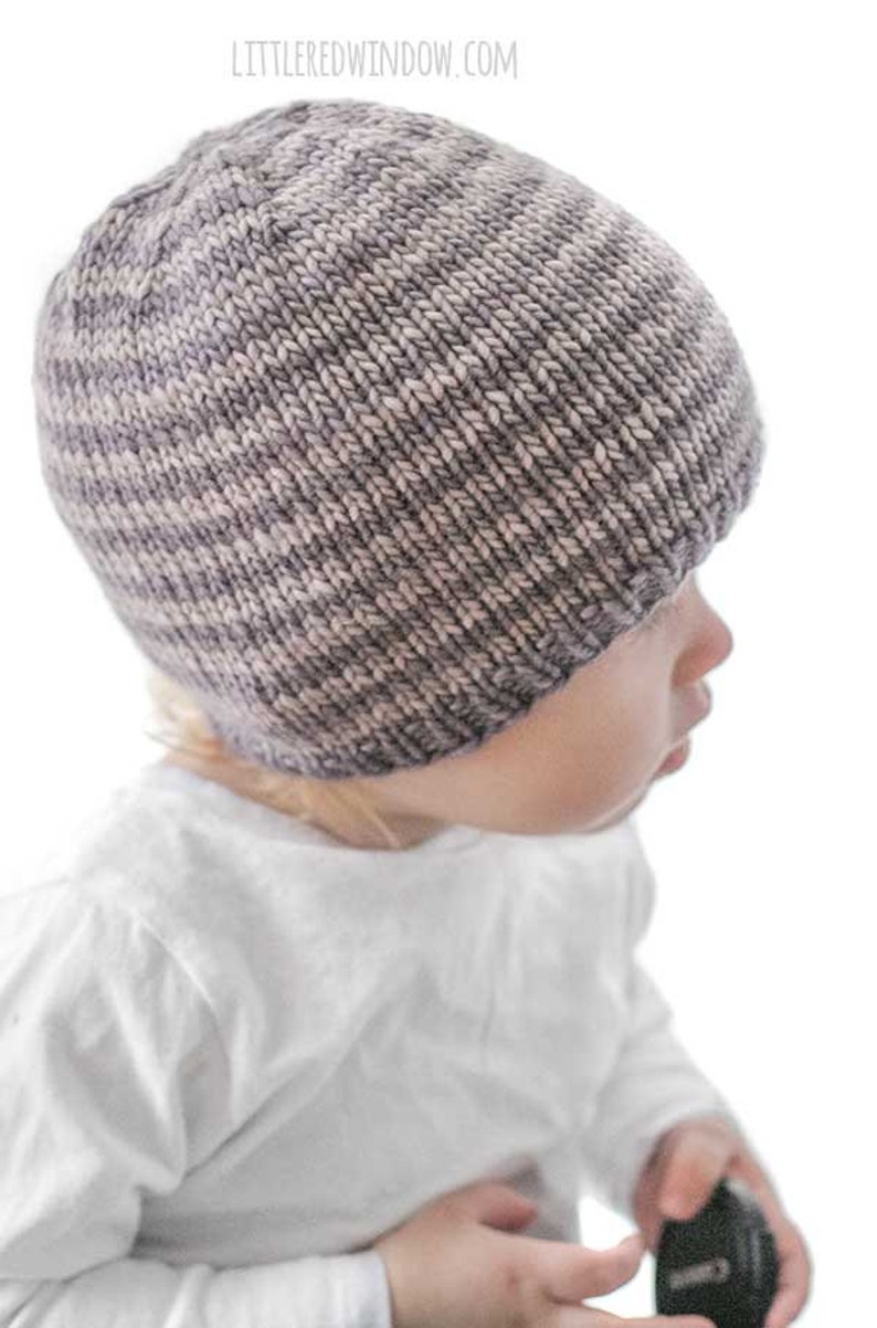 toddler in white shirt wearing a tan and medium brown thin striped knit hat looking off to the right in front of a white background