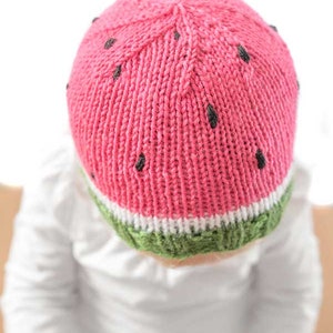 Op de afbeelding: Een roze en groene gebreide watermeloenmuts voor een baby. De muts heeft zwarte pitten en een witte streep.