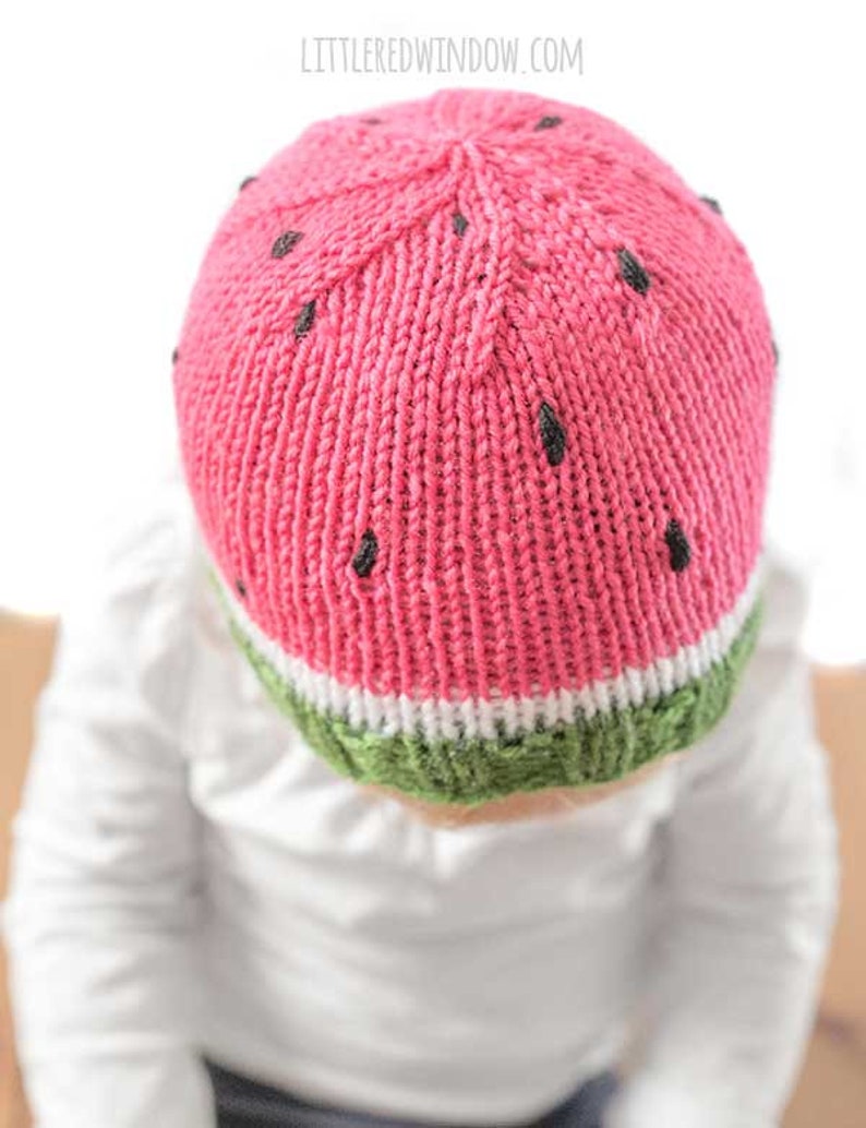 Op de afbeelding: Een roze en groene gebreide watermeloenmuts voor een baby. De muts heeft zwarte pitten en een witte streep.