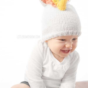 grinning baby in white shirt wearing a white knit hat with golden unicorn horn and rainbow knit mane sitting in front of a white background and looking slightly to the right