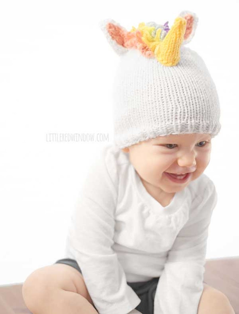 grinning baby in white shirt wearing a white knit hat with golden unicorn horn and rainbow knit mane sitting in front of a white background and looking slightly to the right