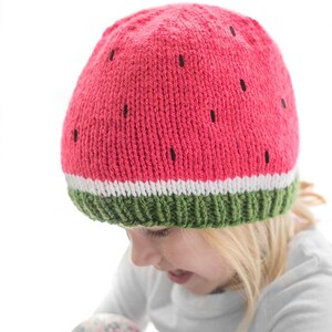 blonde child wearing a knit hat in green white and pink that looks like a watermelon slice looking down to the right