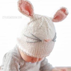 baby in gray shirt wearing a white knit bunny hat with a pink nose gray whiskers and pink lined bunny ears on top looking down at their hands