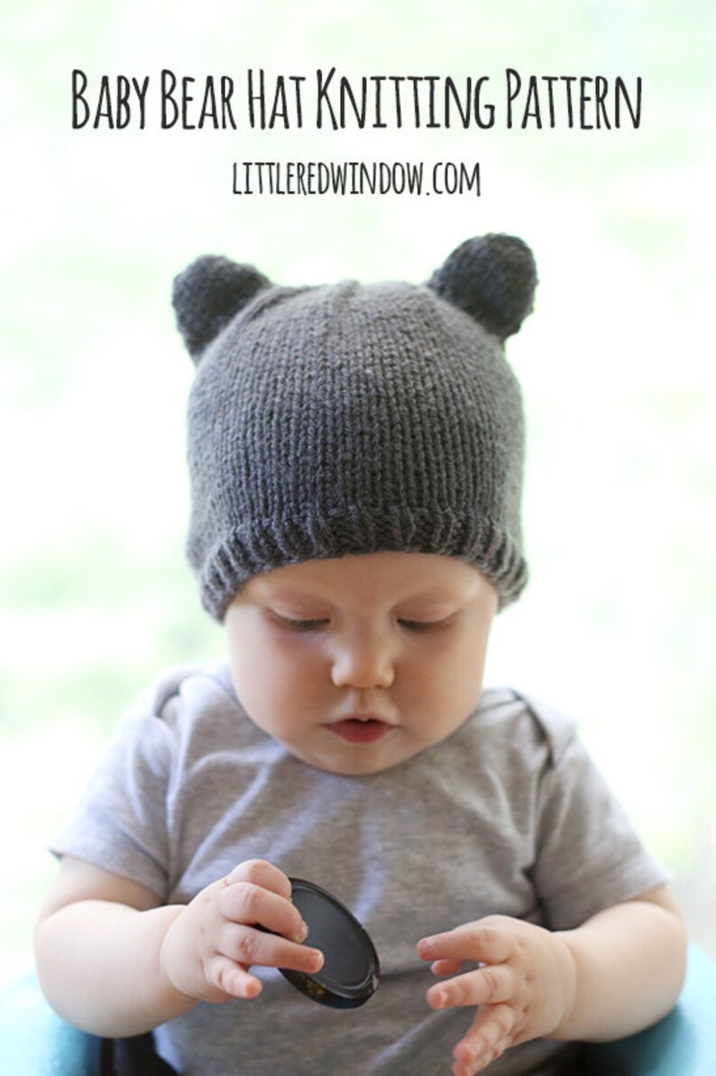 front view of chubby baby in gray short sleeved shirt sitting in a blue chair wearing a dark gray knit hat with bear ears on top and looking down at a camera lens cap in their hands