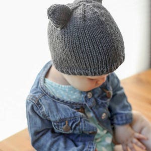 view from above left of baby in light blue onesie and jean jacket sitting on a wood table in front of a white background and wearing a dark gray knit baby hat with bear ears on top