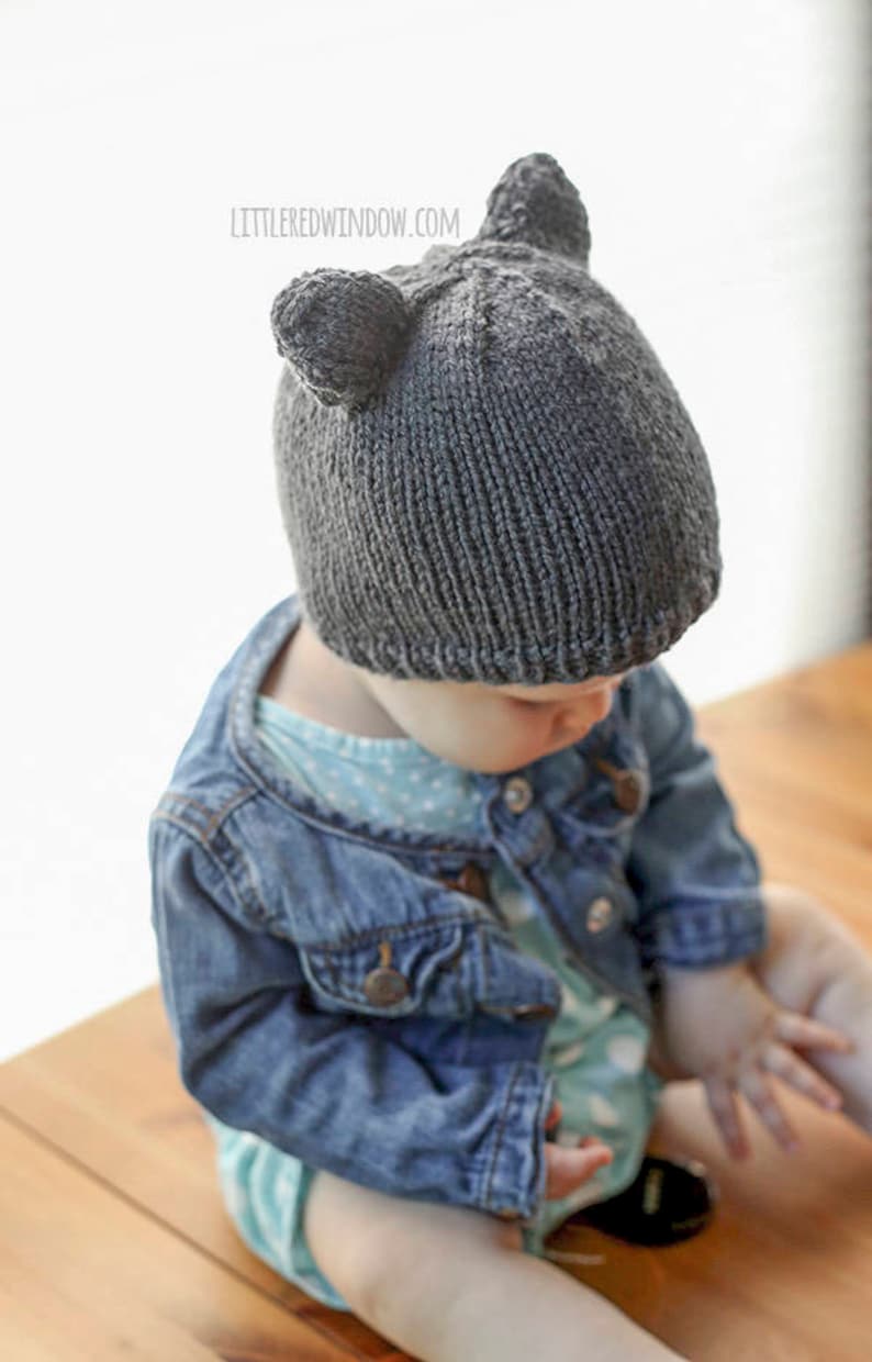 view from above left of baby in light blue onesie and jean jacket sitting on a wood table in front of a white background and wearing a dark gray knit baby hat with bear ears on top