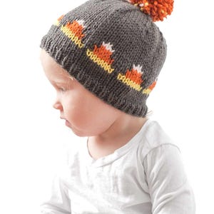 A toddler wearing a gray knitted candy corn hat with orange, white, and yellow accents and an orange pom-pom on top. The child is dressed in a white long-sleeve shirt and is looking downwards. The background is plain white.