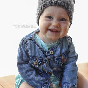 baby sitting on a wood table and smiling with no teeth and wearing a dark gray bear hat with ears and a jean jacket