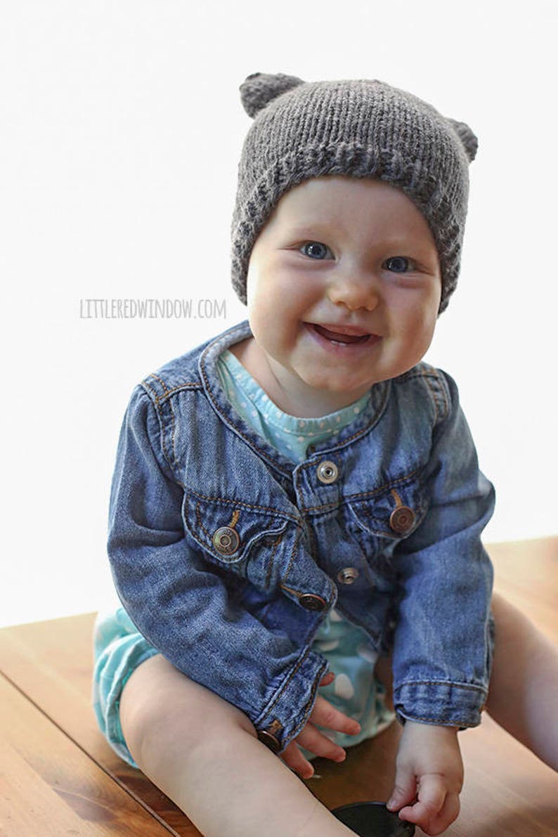 baby sitting on a wood table and smiling with no teeth and wearing a dark gray bear hat with ears and a jean jacket