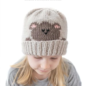 May include: A knitted beige hat featuring a bear face design. The bear has brown fur, pink ears, and a black nose. The hat is worn by a person with blonde hair. Text on the image reads "Peekaboo Bear Hat Knitting Pattern" and "littleredwindow.com".