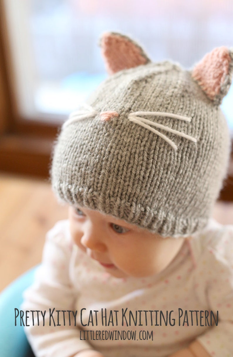 view  of baby in gray onesie wearing a gray knit hat with pink cat ears on top and a little pink nose and white whiskers on the front, looking off to the left while sitting a blue bumbo chair