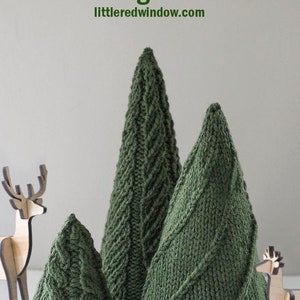 Three dark green knit cone shaped knit Christmas Trees with three different cable knit stitch patterns on a white table with two small wooden deer in front of a gray wall