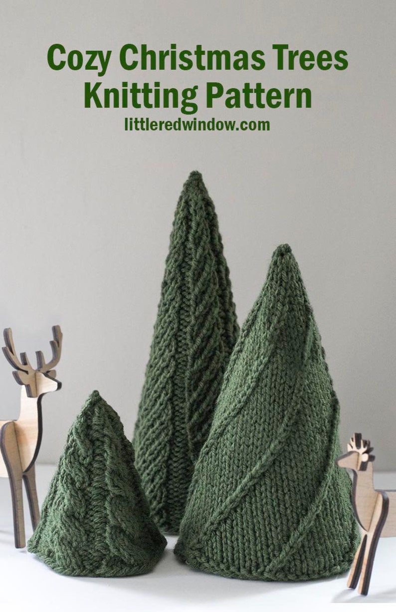 Three dark green knit cone shaped knit Christmas Trees with three different cable knit stitch patterns on a white table with two small wooden deer in front of a gray wall