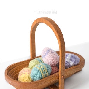 seven pastel colored knit Easter eggs piled in a medium brown low wood basket with a wood handle on a white tabletop