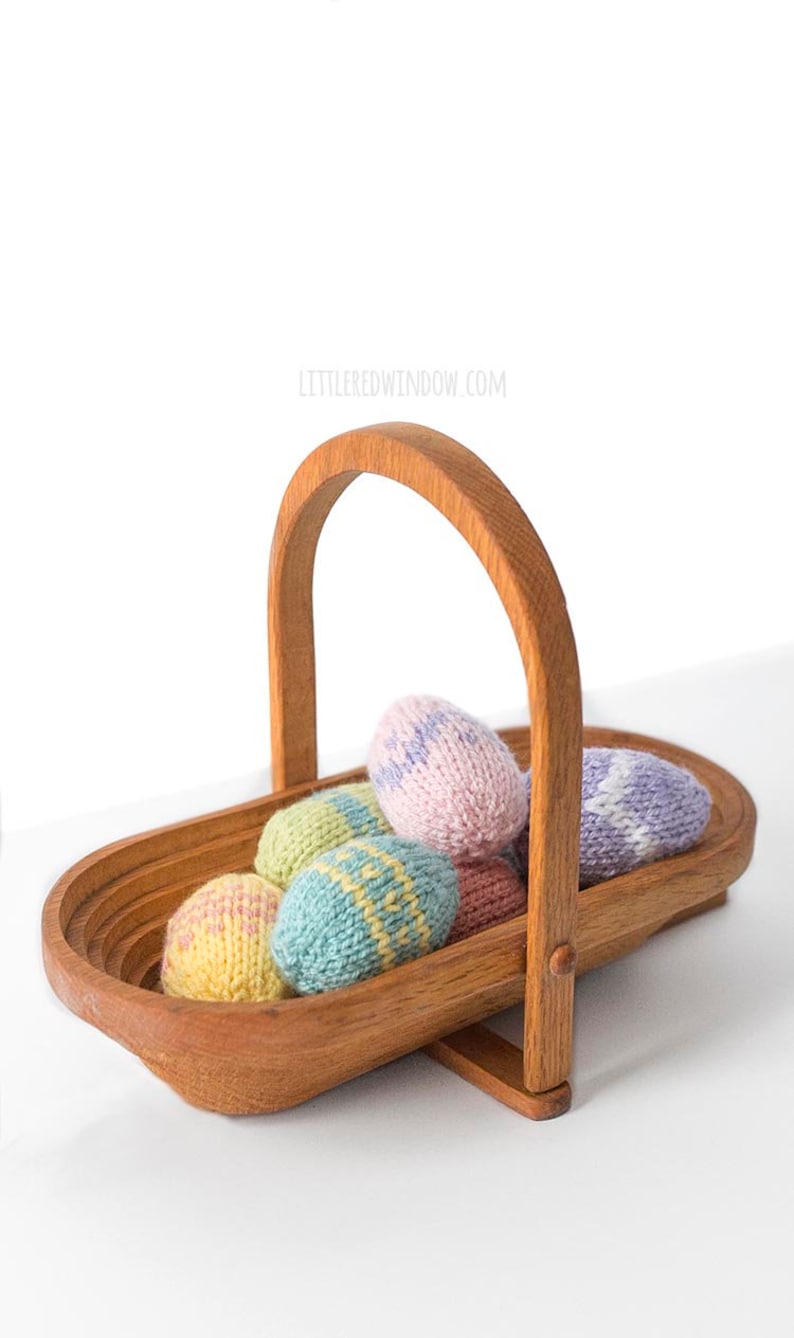 seven pastel colored knit Easter eggs piled in a medium brown low wood basket with a wood handle on a white tabletop