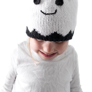 K&ouml;nnte beinhalten: Ein Kind tr&auml;gt eine wei&szlig;e gestrickte Geisterm&uuml;tze mit schwarzen Augen und einem L&auml;cheln. Die M&uuml;tze hat einen schwarzen gewellten Rand.