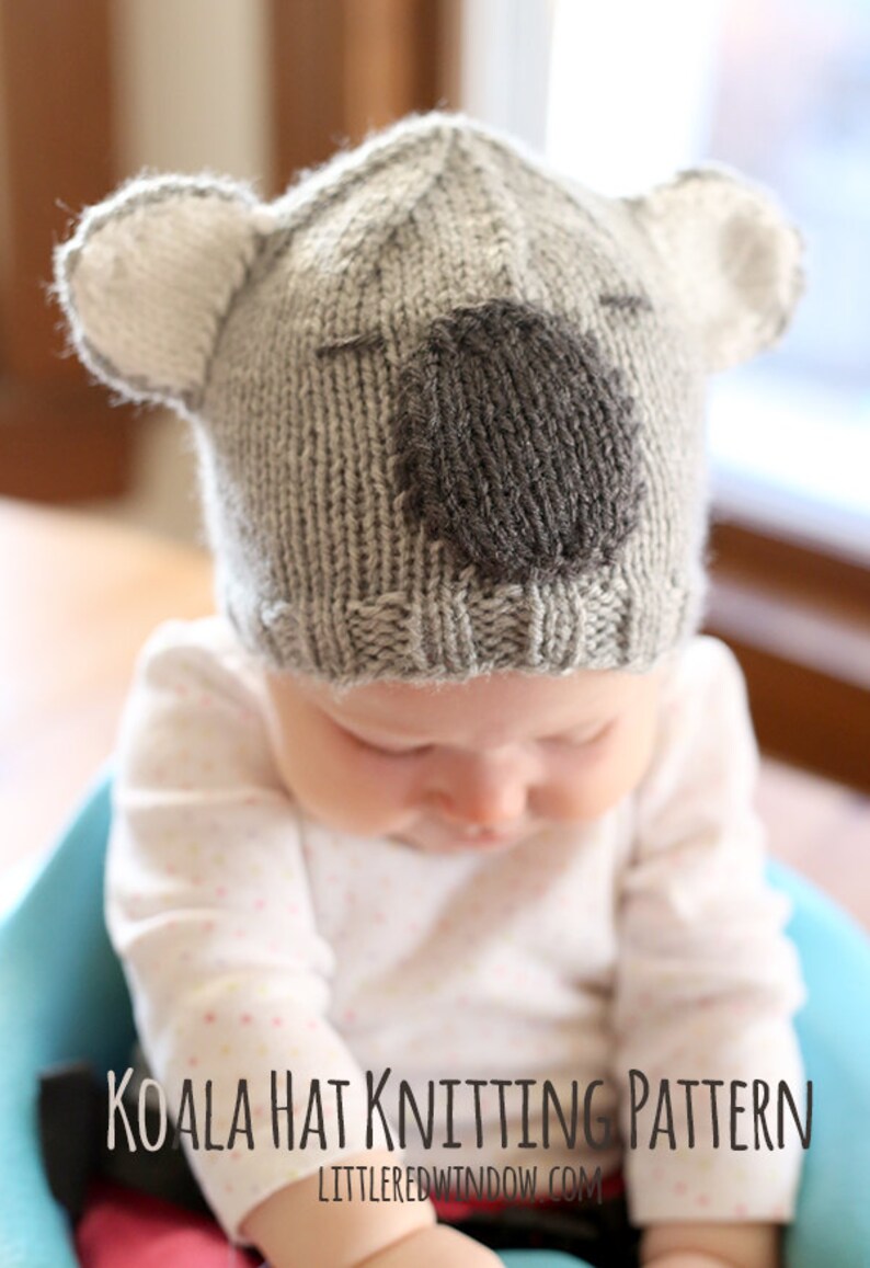 Baby in a pink and white shirt sitting in a blue chair wearing a gray knit hat that looks like a koala looking down and to the right