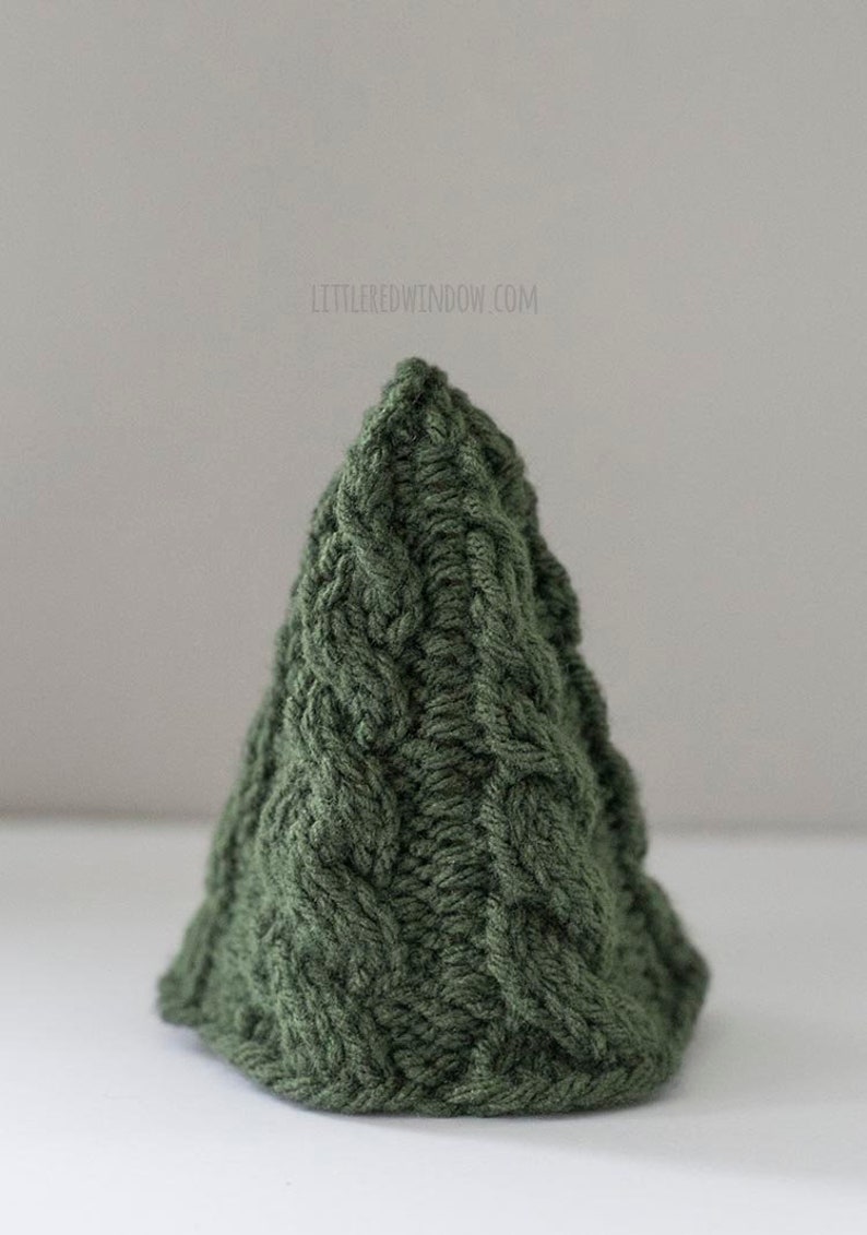 The small Cozy Christmas tree knitting pattern features a simple twisted cable pattern in a tree shape knit in dark green yarn sitting on a white table in front of a gray wall