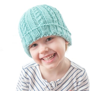 May include: A child wearing a light blue knitted hat with a ribbed pattern. The hat is pulled down over the child's forehead. The child is smiling and wearing a white and blue striped shirt with a small red heart on the front. The text "Alternating Rib Hat Knitting Pattern" and "littleredwindow.com" is visible at the top of the image.