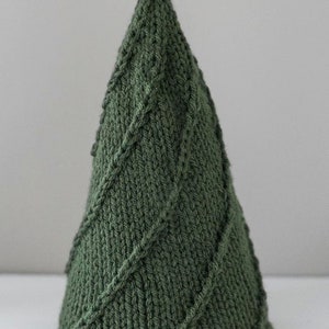 The mediun Cozy Christmas tree knitting pattern features  simple lines of twisted stitches in a tree shape knit in dark green yarn sitting on a white table in front of a gray wall