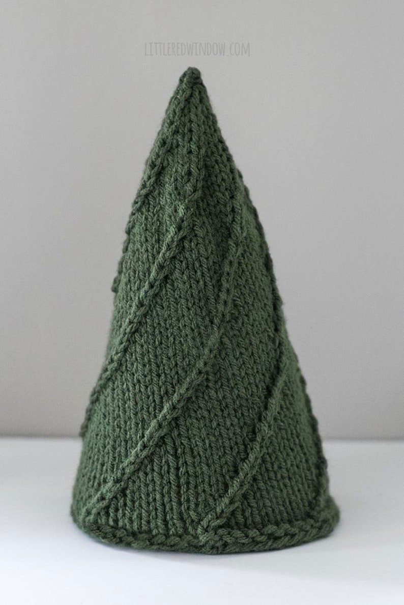 The mediun Cozy Christmas tree knitting pattern features  simple lines of twisted stitches in a tree shape knit in dark green yarn sitting on a white table in front of a gray wall