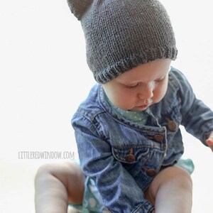 Baby in a jean jacket reaching for a lens cap on the table and wearing a dark gray knit hat with bear ears on top