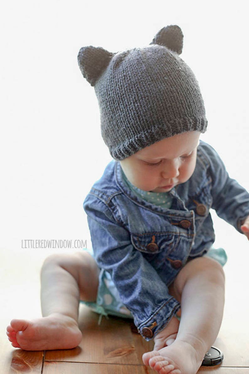 Baby in a jean jacket reaching for a lens cap on the table and wearing a dark gray knit hat with bear ears on top