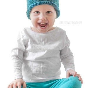 toddler in white shirt and blue pants wearing a knit hat with a rolled brim made of teal blue hand dyed yarn  sitting on a wood table and smiling at the camera