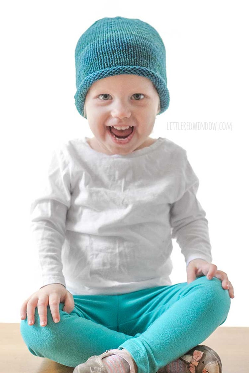 toddler in white shirt and blue pants wearing a knit hat with a rolled brim made of teal blue hand dyed yarn  sitting on a wood table and smiling at the camera