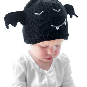 A baby in a white long-sleeve shirt wears a Bat Hat, a black knit cap designed to resemble a bat with wings and a face featuring stitched eyes, a smile, and tiny fangs. The baby is looking down against a plain white background.