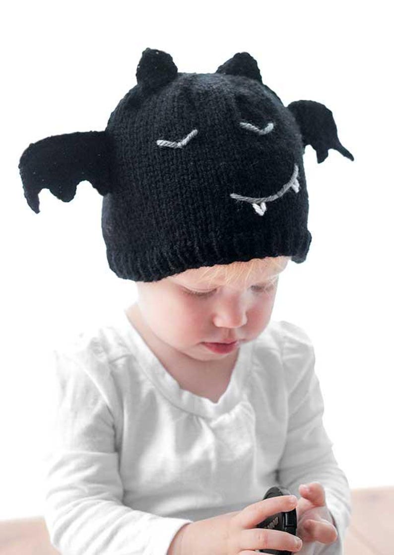 A baby in a white long-sleeve shirt wears a Bat Hat, a black knit cap designed to resemble a bat with wings and a face featuring stitched eyes, a smile, and tiny fangs. The baby is looking down against a plain white background.