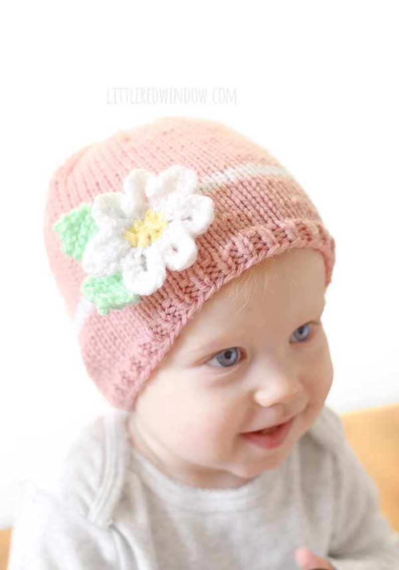 closeup of baby wearing light pink knit hat with white knit band and white knit flower on the front smiling and looking to the right
