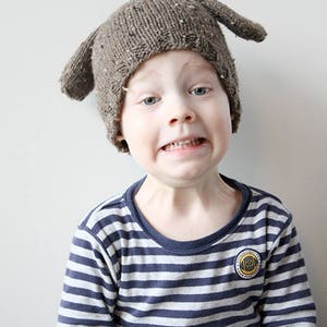 Puede incluir: Un ni&ntilde;o lleva un gorro de punto marr&oacute;n de cachorro con orejas ca&iacute;das. El gorro tiene un patr&oacute;n de punto texturizado. El ni&ntilde;o lleva una camisa de manga larga a rayas azules y blancas.