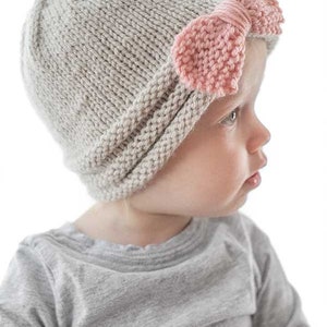girl in gray shirt wearing tan knit hat gathered in the front with large pink knit bow looking down off to the right in front of a white background