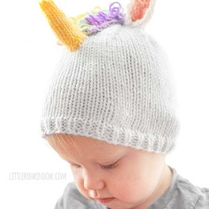serious toddler in gray shirt wearing a white knit hat with a unicorn horn and rainbow mane on top looking down at something in their hands
