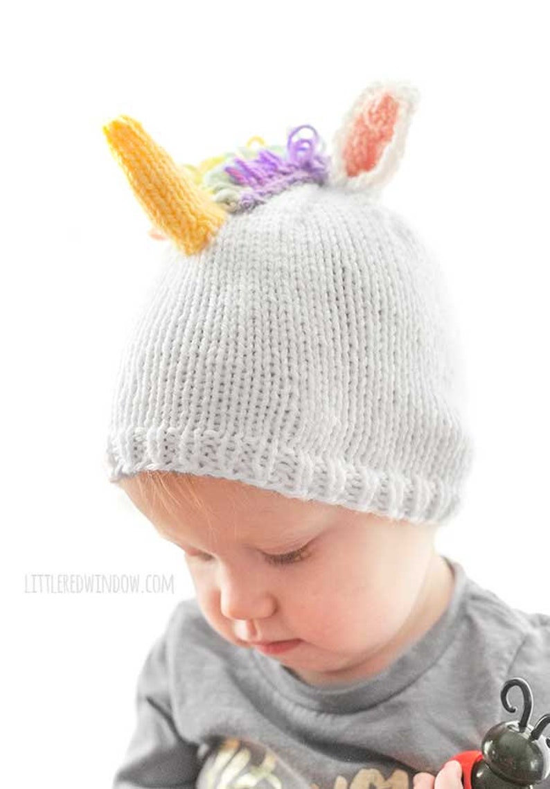 serious toddler in gray shirt wearing a white knit hat with a unicorn horn and rainbow mane on top looking down at something in their hands