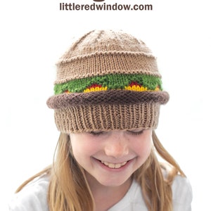 May include: A brown knitted hat shaped like a hamburger with green lettuce, red tomato, yellow cheese, and a brown bun. The hat is being worn by a young person with blonde hair.