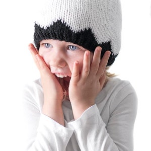 K&ouml;nnte beinhalten: Ein Kind tr&auml;gt eine wei&szlig;e und schwarze Strickm&uuml;tze mit einem l&auml;chelnden Gesicht und schwarzen Augen. Das Kind h&auml;lt seine H&auml;nde an seine Wangen und hat einen &uuml;berraschten Gesichtsausdruck.