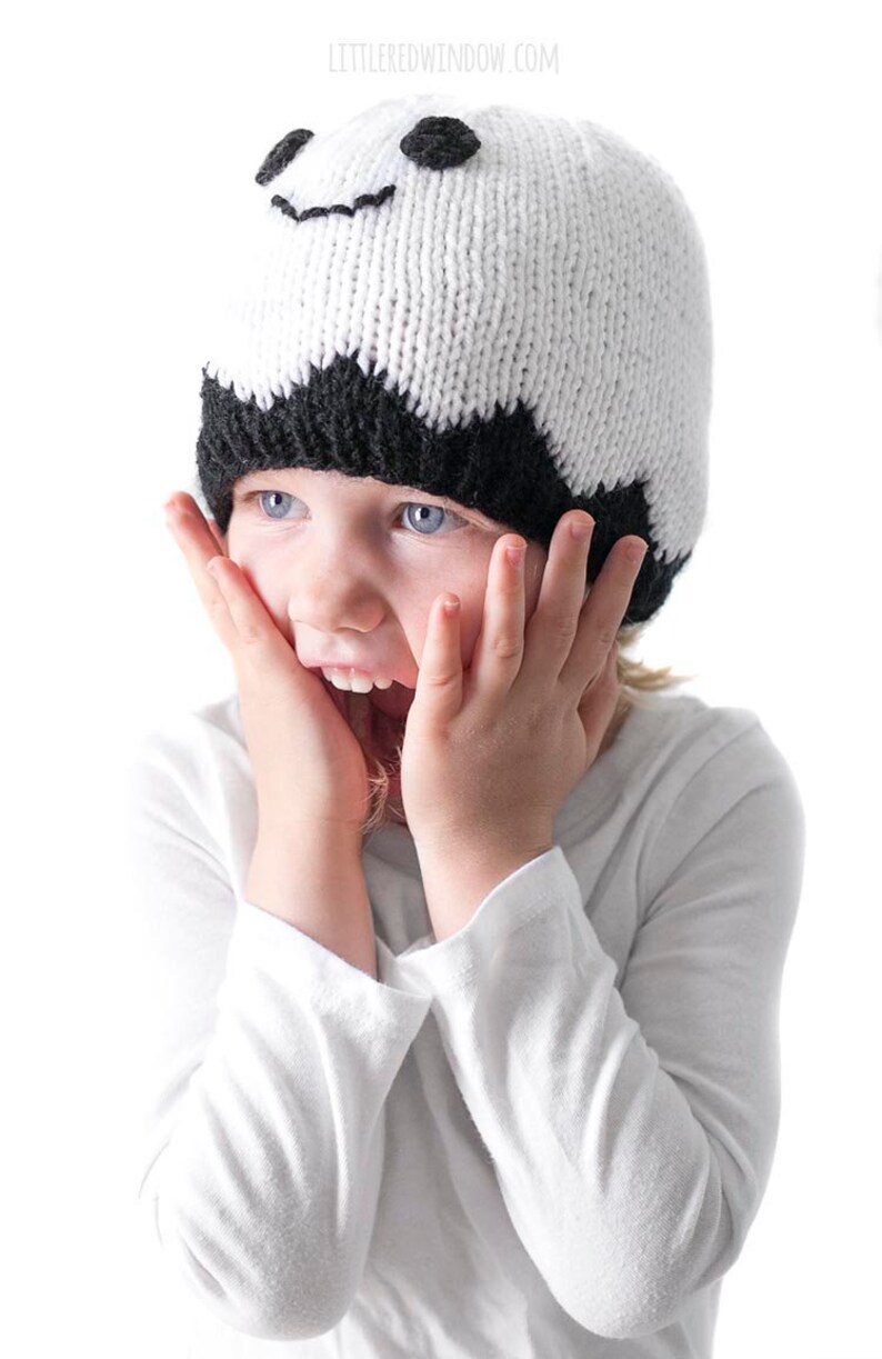K&ouml;nnte beinhalten: Ein Kind tr&auml;gt eine wei&szlig;e und schwarze Strickm&uuml;tze mit einem l&auml;chelnden Gesicht und schwarzen Augen. Das Kind h&auml;lt seine H&auml;nde an seine Wangen und hat einen &uuml;berraschten Gesichtsausdruck.