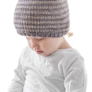 toddler in white shirt wearing a tan and medium brown thin striped knit hat looking down at their lap in front of a white background