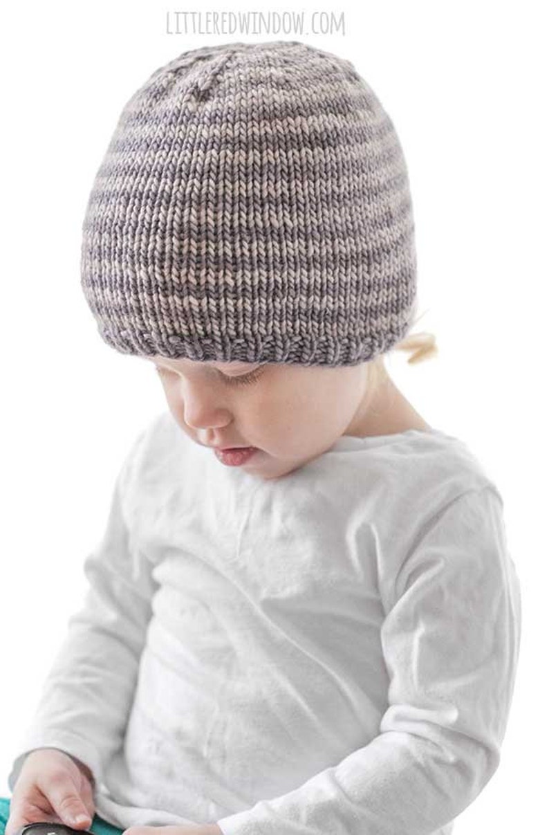 toddler in white shirt wearing a tan and medium brown thin striped knit hat looking down at their lap in front of a white background