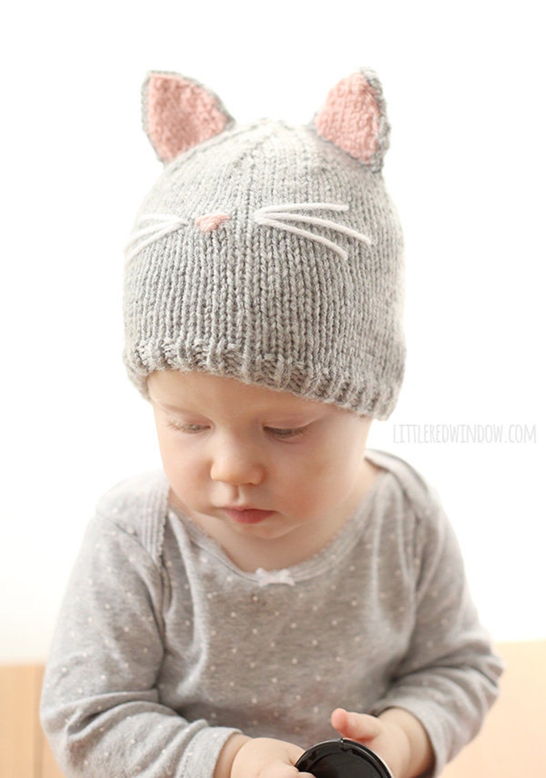 baby in gray onesie wearing a gray knit hat with pink cat ears on top and a little pink nose and white whiskers on the front, looking down and to the left and holding a camera lens cap in their hand