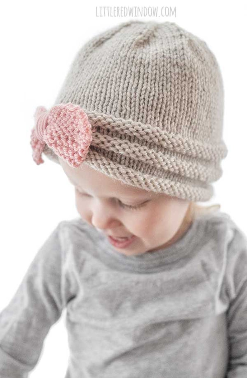 view from slightly above of smiling girl in gray shirt wearing tan knit hat gathered in the front with large pink knit bow looking down to the right
