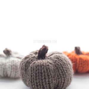three small knit pumpkins knit with orange tan and medium brown yarn with dark brown knit pumpkin stems sitting on a white table with the brown pumpkin in front slightly taller than the other two