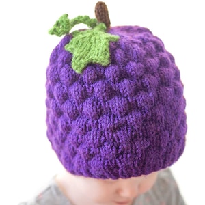 Könnte beinhalten: Eine violette gestrickte Traubenmütze mit grünem Stiel und Blatt. Die Mütze wird von einem Kind mit einem grauen Shirt getragen.