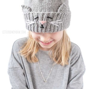 May include: A gray knitted hat with a cat face design. The hat has white whiskers and a pink nose. The hat is being worn by a child with blonde hair.