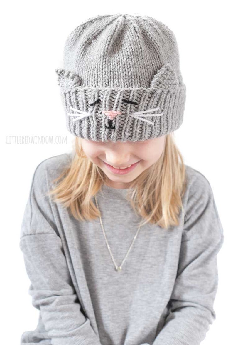 May include: A gray knitted hat with a cat face design. The hat has white whiskers and a pink nose. The hat is being worn by a child with blonde hair.
