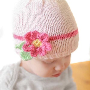 closeup of baby in gray onesie wearing a light pink knit hat with hot pink band and flower around the middle looking down to the right