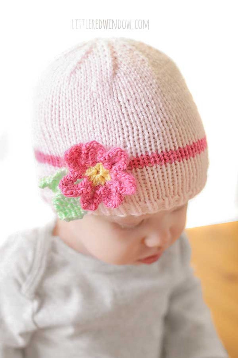 closeup of baby in gray onesie wearing a light pink knit hat with hot pink band and flower around the middle looking down to the right