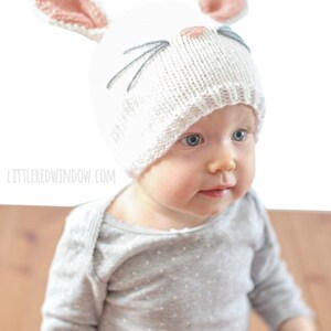 baby in gray shirt wearing a white knit bunny hat with a pink nose gray whiskers and pink lined bunny ears on top looking down at their hands  looking up and off to the right